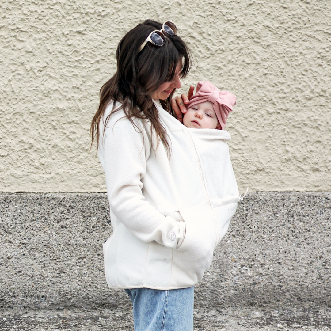 Mother carrying baby under light fleece babywearing jacket, both cuddled closely