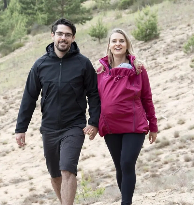 Ein junger Mann mit schwarzer Jacke und eine junge Frau mit violeter Tragejacke und Baby an der Brust wandern Hand in Hand eine karge Grasfläche entlang.