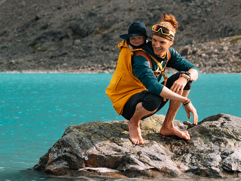 Ein junger Mann hockt auf einem großen Stein in einem Gebirgssee. Auf den Rücken trägt er eine orangene mamalila Tragejacke mit Baby.