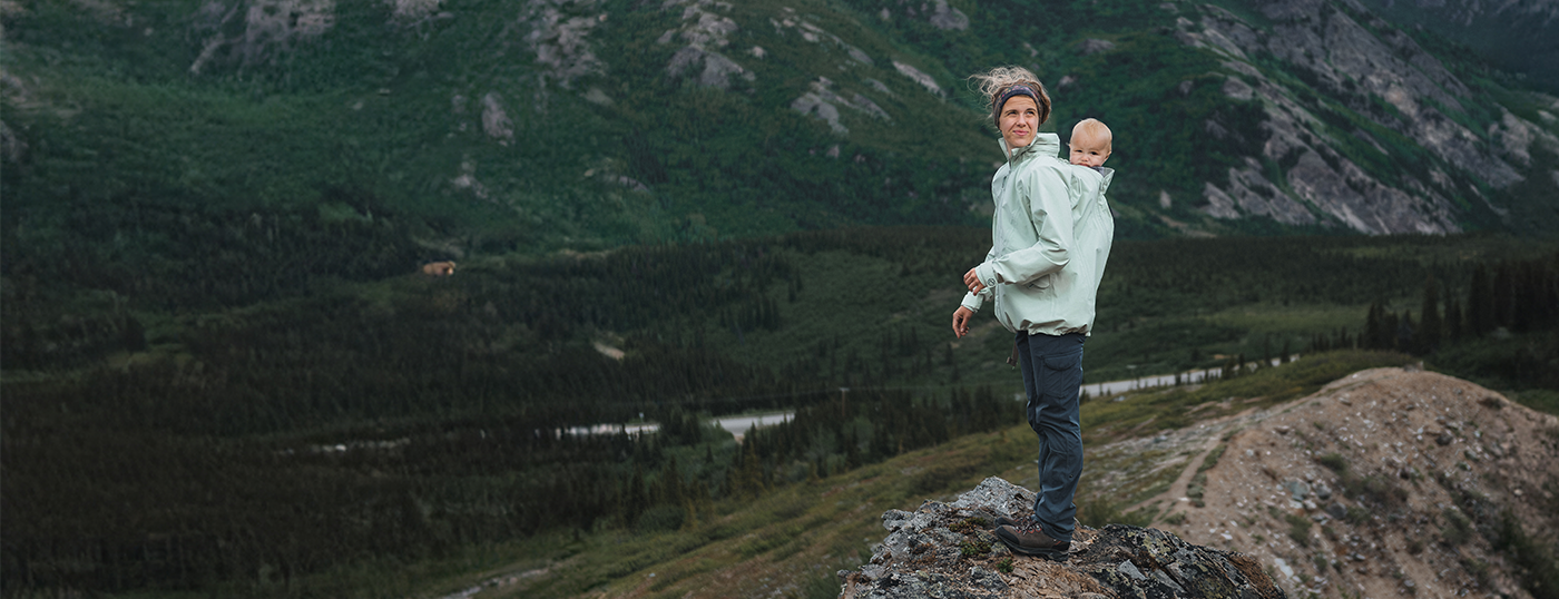 Mother carrying baby on her back in light babywearing jacket, standing on mountain ridge