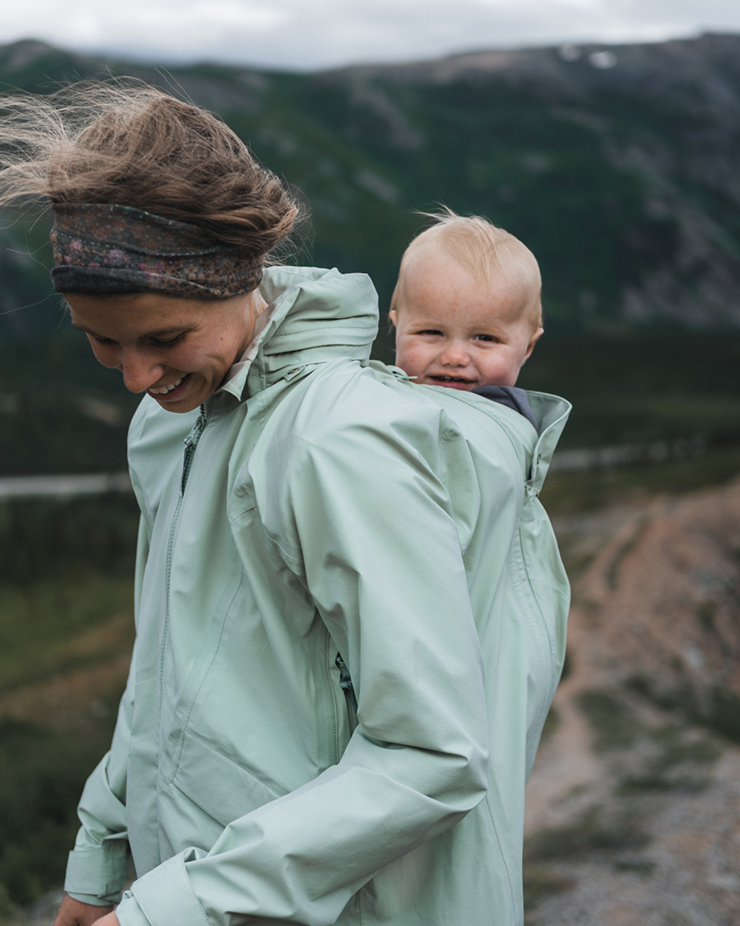 Mother carrying baby on her back in green babywearing jacket, hiking in mountainous landscape