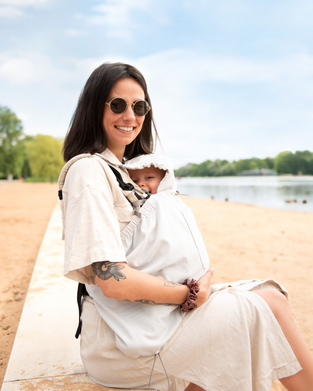 Mother sitting on beach carrying baby under light babywearing cover, sunny scene