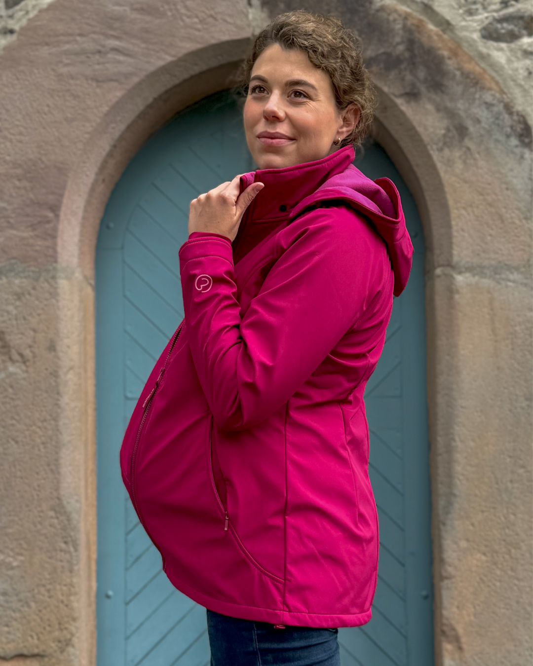 Eine schwangere Frau trägt eine pinke Softshell-Tragejacke und steht vor einer Tür.