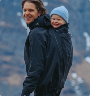 Father carrying baby on his back in dark babywearing jacket, both smiling outdoors