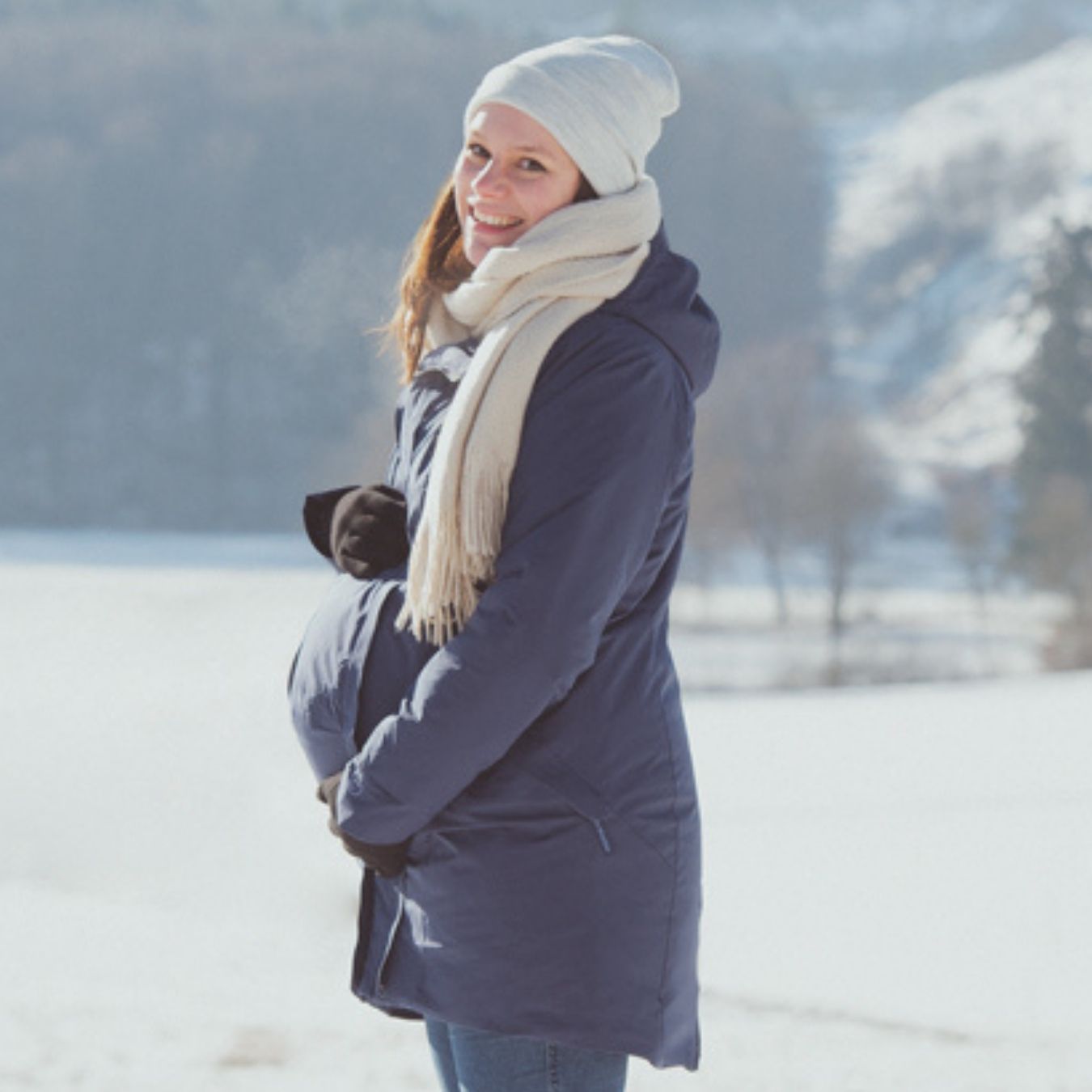 Pregnant woman in blue winter jacket with hat outdoors in snowy landscape