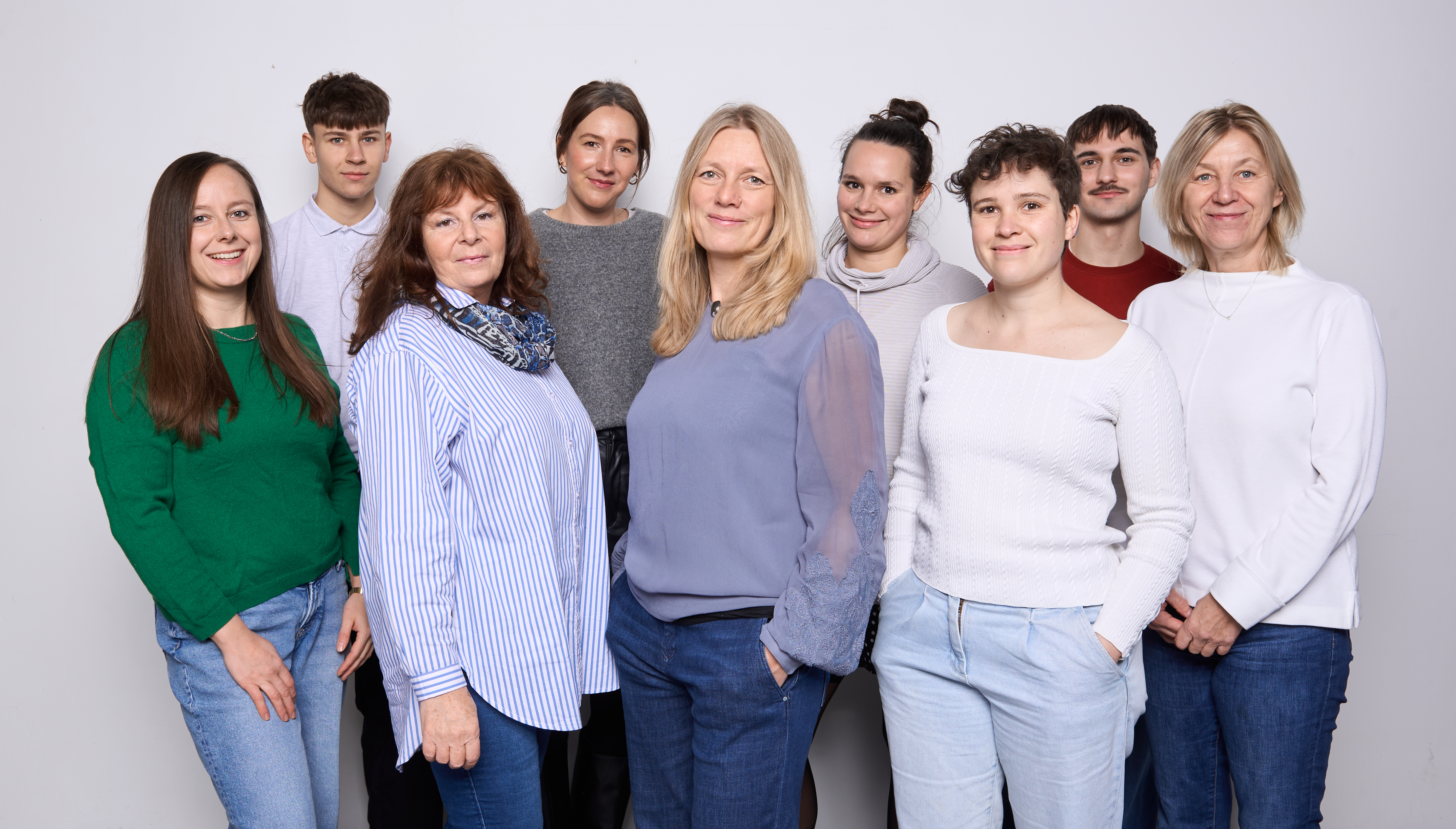 Group of nine people standing together against light background, smiling at camera