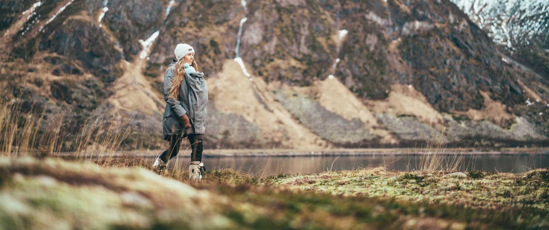 Mother carrying baby in babywearing jacket by lakeside with mountains, outdoor setting