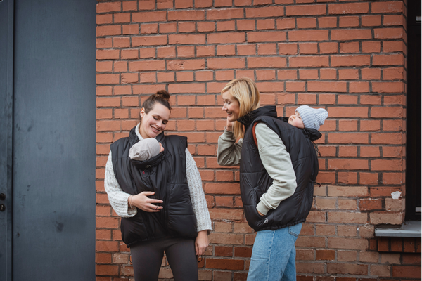 Two women with babies in carriers wearing black vests in front of brick wall
