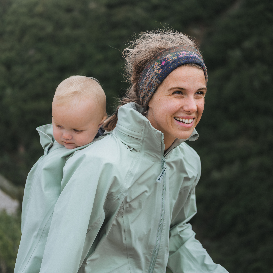Woman carrying baby on back in light jacket, smiling outdoors
