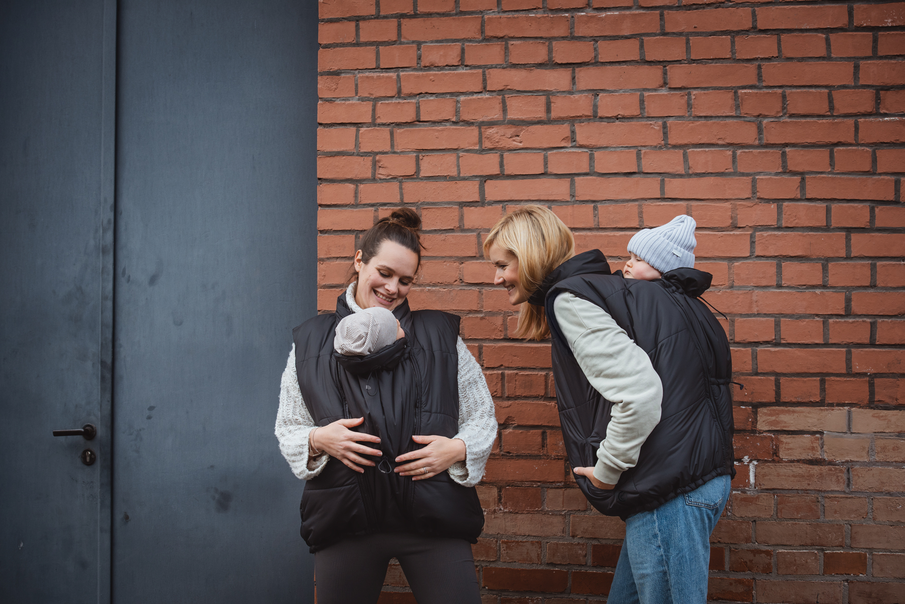 Two mothers carrying babies front and back with black vests in front of brick wall