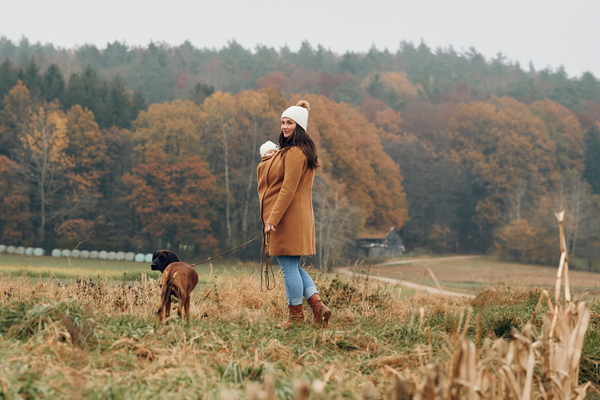 Woman with baby in carrier coat standing in field, dog on leash, autumn scenery