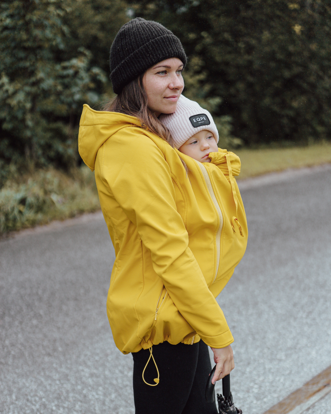 Woman carrying baby under yellow jacket, standing outdoors on wet road