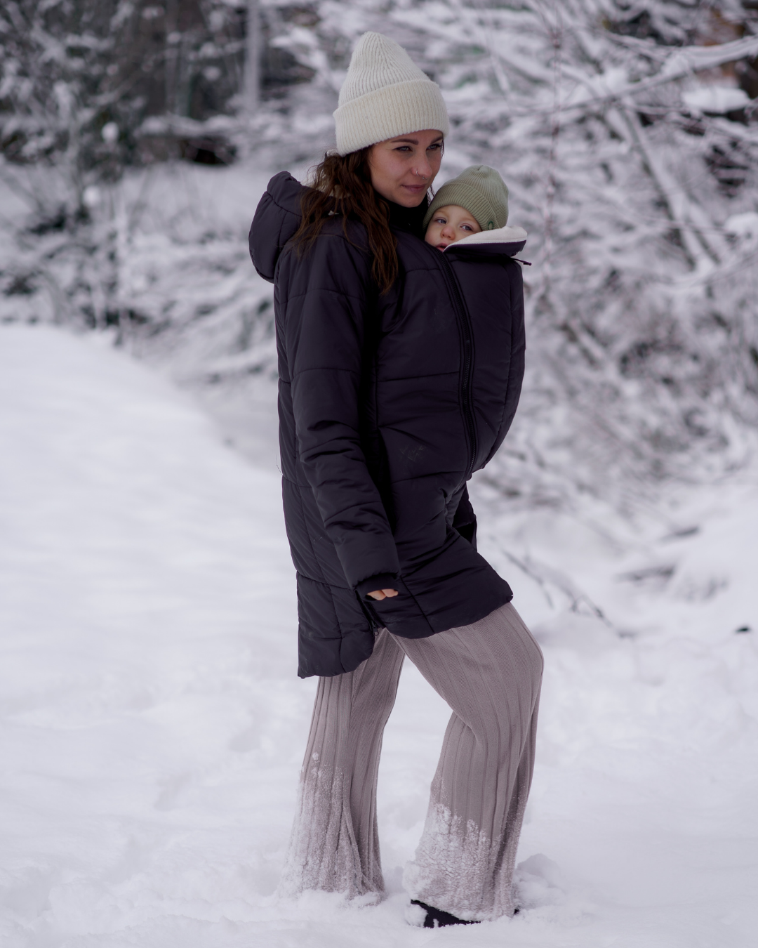 Mother carrying baby in dark quilted babywearing jacket in snow, winter landscape, warmly dressed