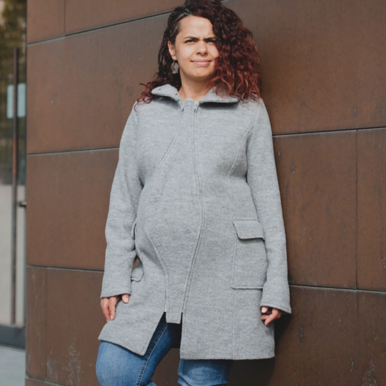 Pregnant woman in grey jacket standing by wall, visible baby bumpPregnant woman in grey jacket standing by wall, visible baby bump