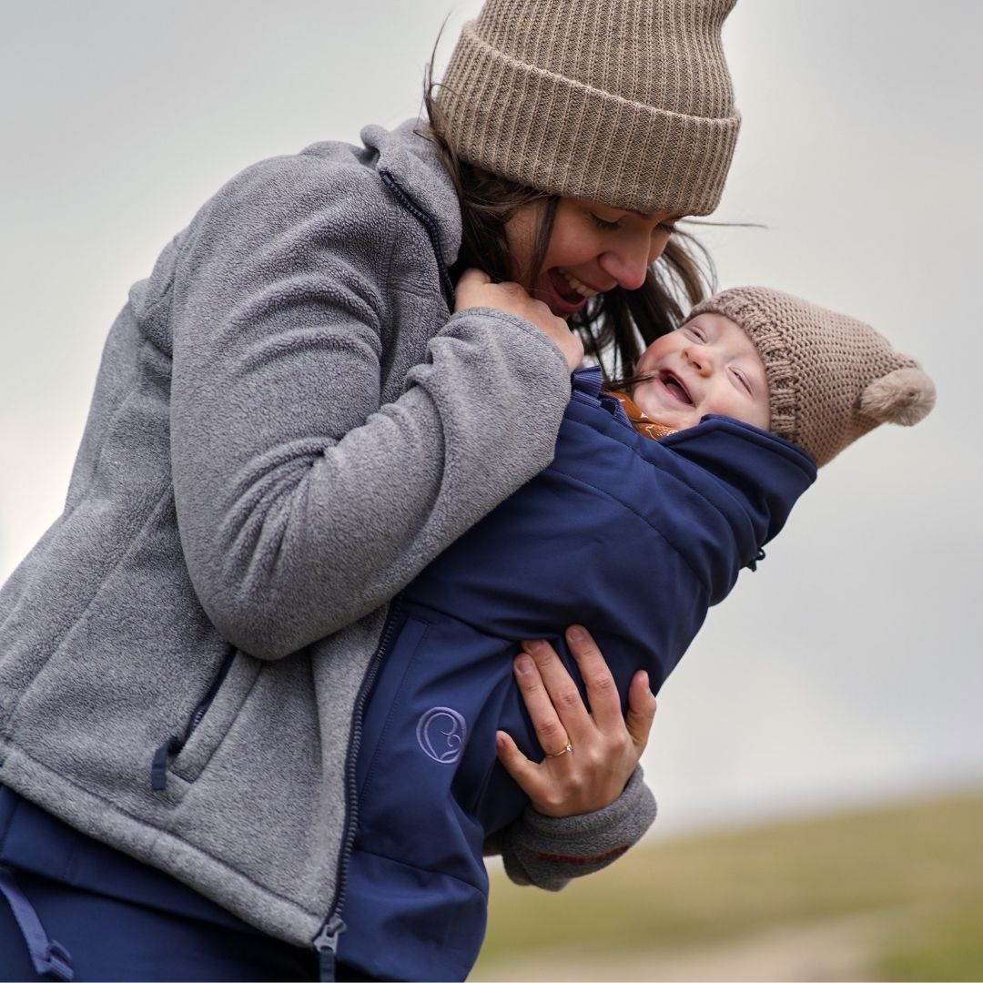 Mother holding baby in carrier cover, both wearing hats and smiling