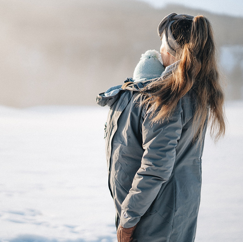 Mother carrying baby in grey babywearing jacket in snow, looking into calm winter landscape