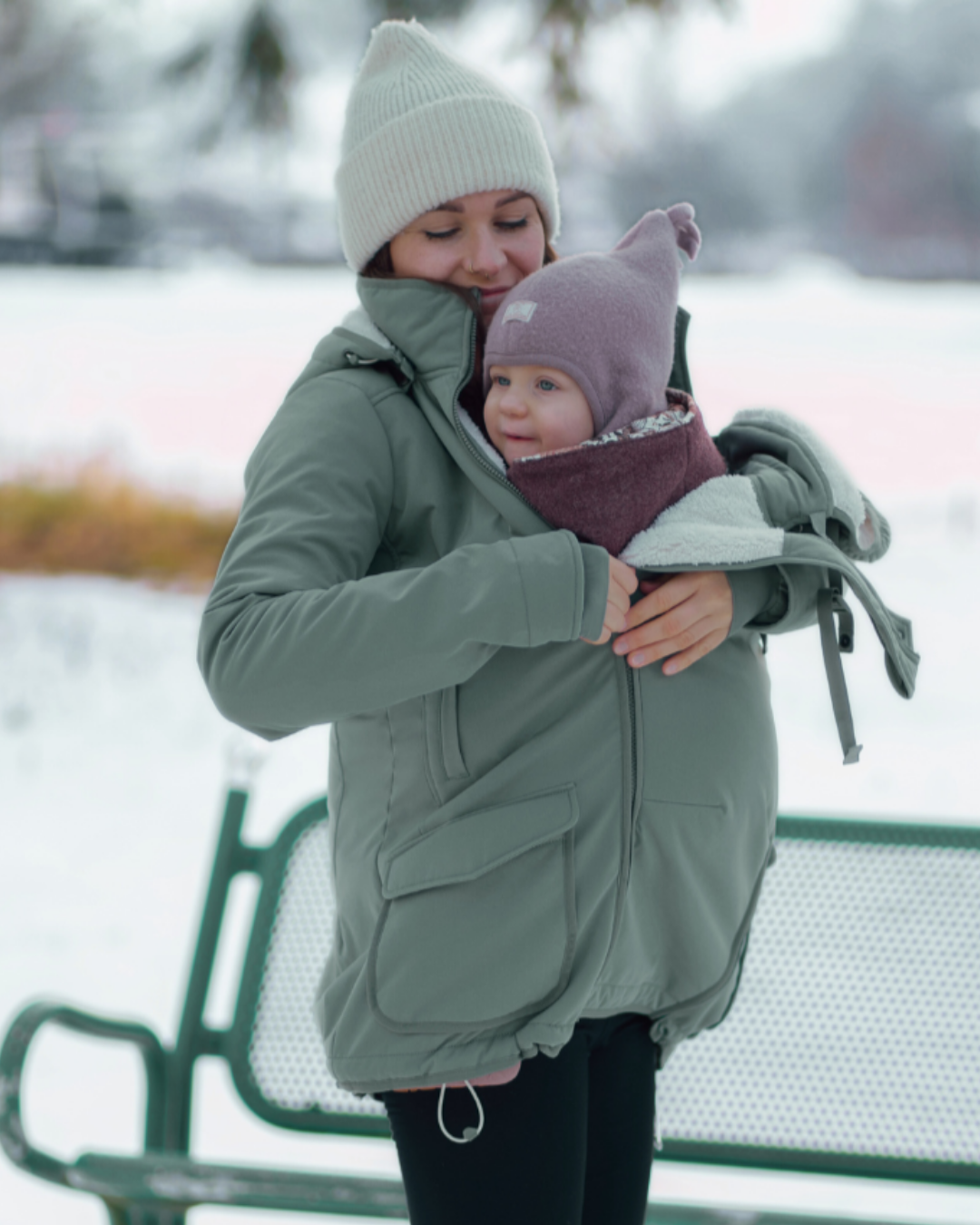 Mother carrying baby in green babywearing jacket in snow, both wearing hats