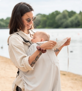 Mother carrying baby in light babywearing cover by the lake, sitting and reading