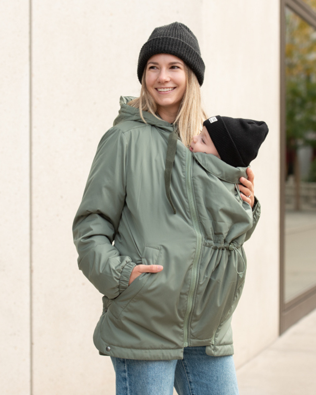 Mother carrying baby in green babywearing jacket, urban autumn scene, both wearing black hats